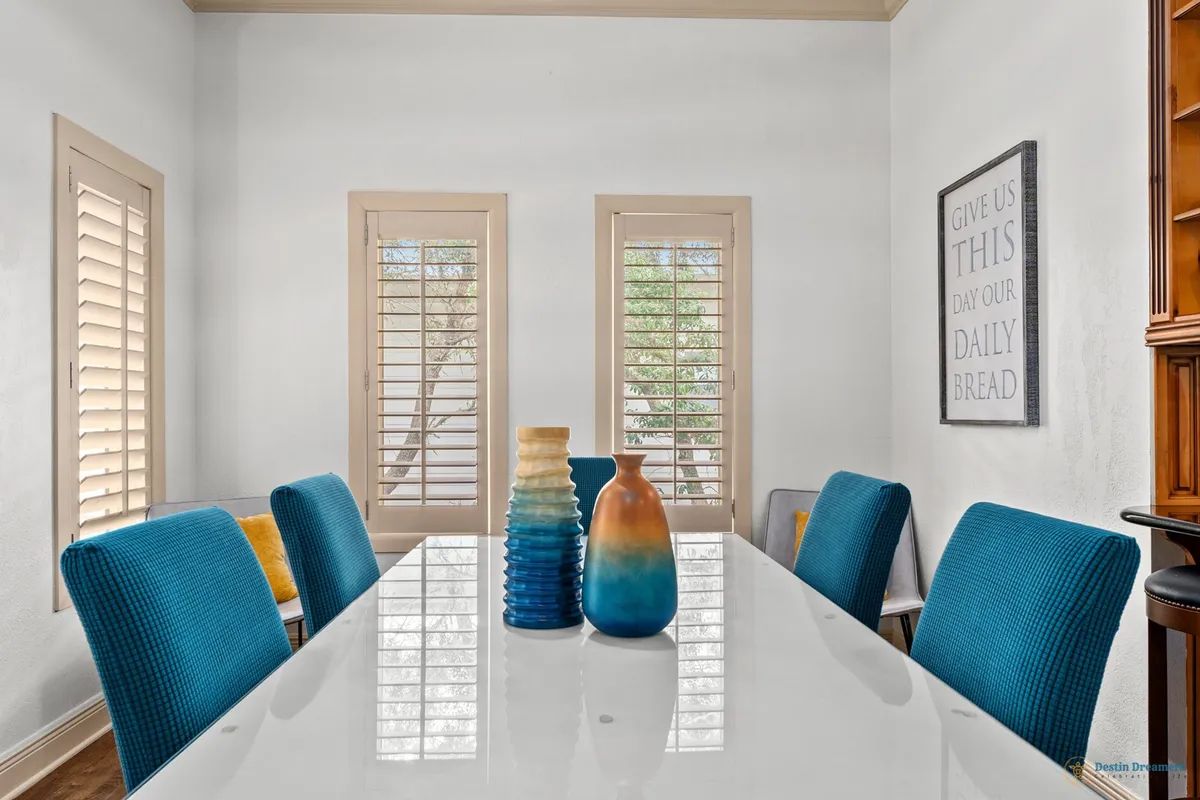 A cheerful dining area illuminated by natural light, with a central table surrounded by blue chairs, colorful vases as centerpieces, and shuttered windows in the background. The ambiance is vibrant and social, perfect for gathering before or after outdoor adventures.