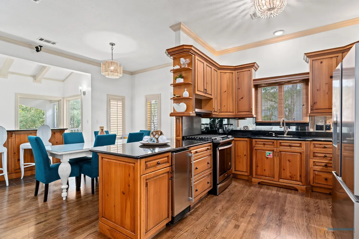 A stylish kitchen with a large central island, blue dining chairs tucked around a wooden dining table, and elegant light fixtures overhead. The kitchen features modern appliances and warm wood cabinetry, creating a cozy yet functional hub for group cooking and meals.