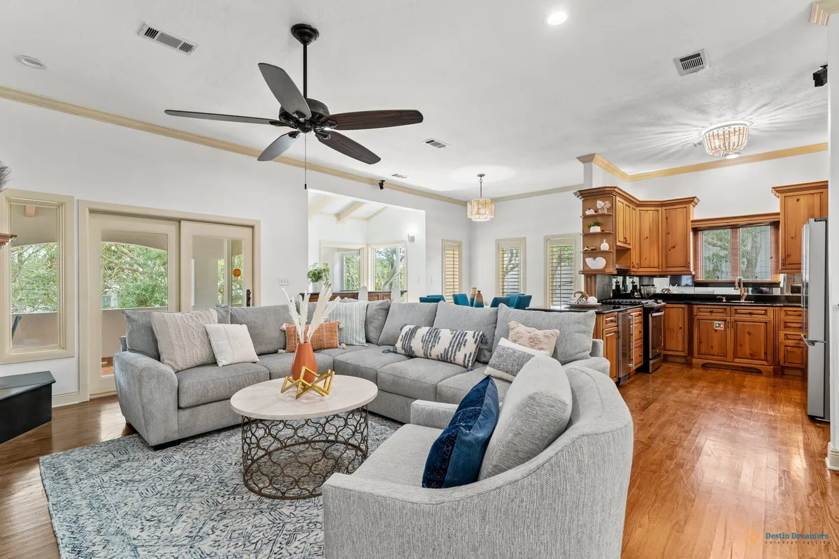 A wide-angle view of an open-plan living area featuring a large sectional sofa and a decorative coffee table, with the adjacent kitchen area visible in the background. The space is bathed in natural light thanks to big windows, fostering a sense of warmth and togetherness.