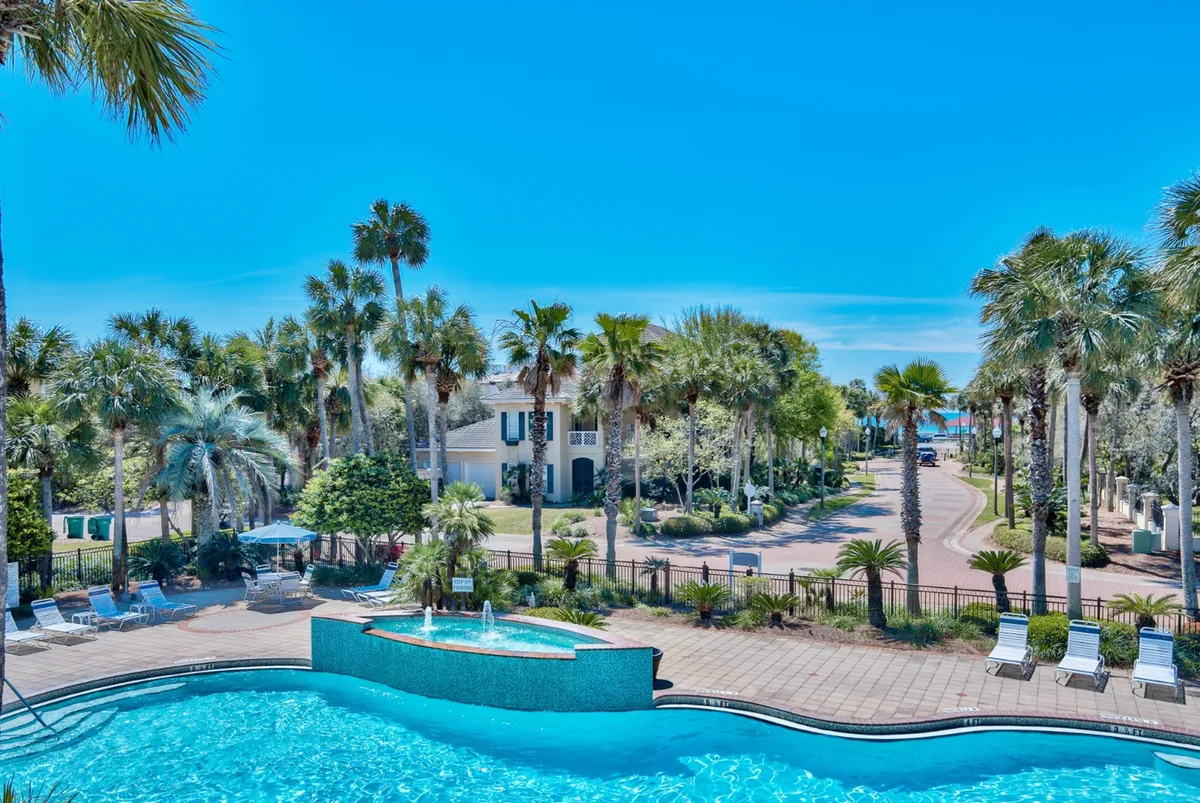 The hero image showcases a sparkling heated community pool with a stunning waterfall edge, framed by lush palm trees and white lounge chairs. A gated pathway leads directly toward the nearby beach, suggesting effortless access to sand and surf. The atmosphere is serene and exclusive, inviting guests to relax poolside or stroll to the ocean within moments.