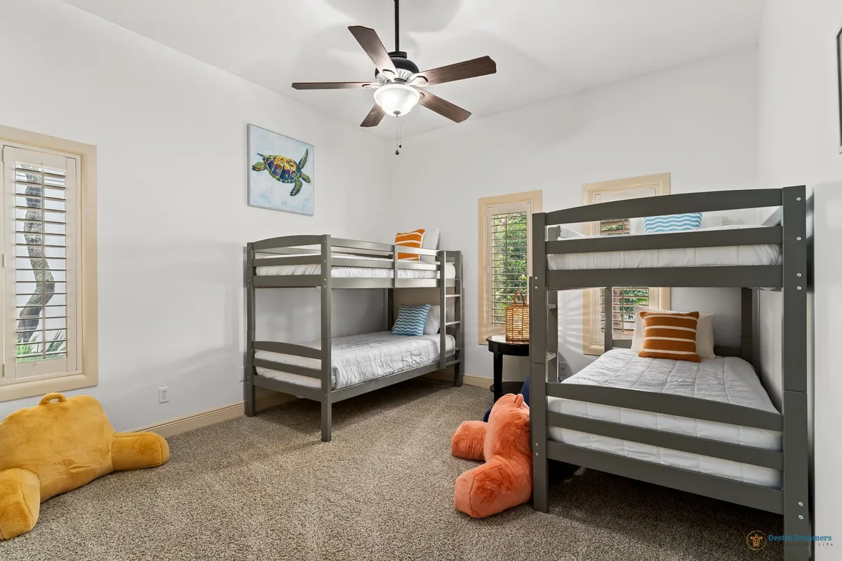 A cheerful bunk and gaming room with two sets of gray bunk beds, white bedding, colorful pillows, plush stuffed animals, plantation shutter windows allowing natural light, and a small round table with a lamp. The space feels playful, comfortable, and family-friendly.