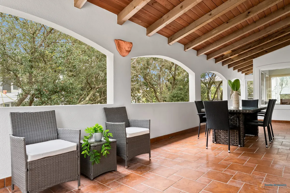 A bright, breezy covered veranda features cushioned wicker armchairs and a dark wicker dining table for six, all set beneath a wooden ceiling with exposed beams. Arched openings frame views of lush gardens, and the area is perfect for relaxing, reading, or enjoying a meal surrounded by nature.