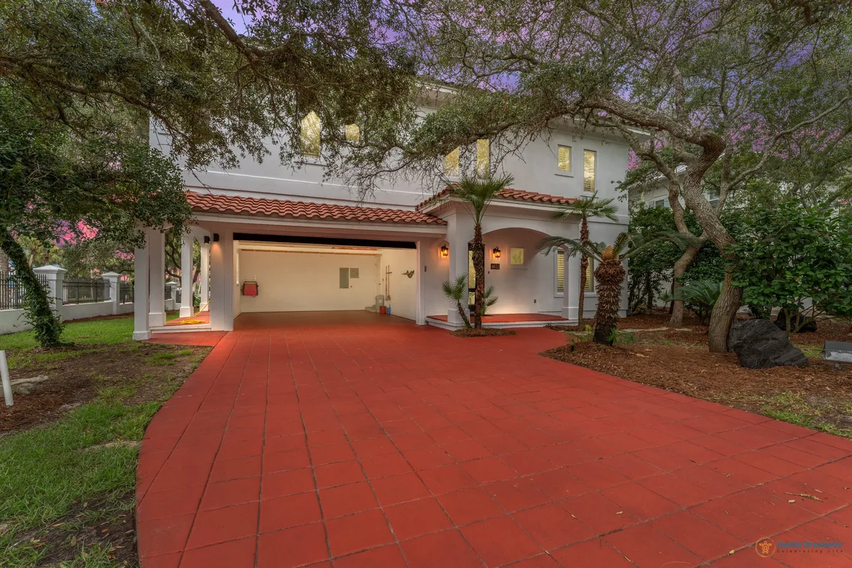 This exterior shot features a large red tiled driveway leading to an open garage with warm lighting, set against a white two-story house with terracotta roof tiles and mature trees framing the property. The scene conveys a sense of arrival and welcoming ambiance, with the home situated in a peaceful, lush environment.