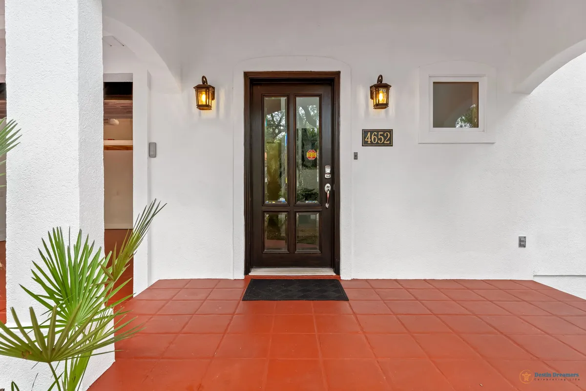 An exterior view of the home’s entrance, showing a dark wooden front door with glass panels, flanked by warm lanterns, bright white stucco walls, and a terracotta tiled floor. The inviting entry sets the tone for a welcoming, luxurious stay.