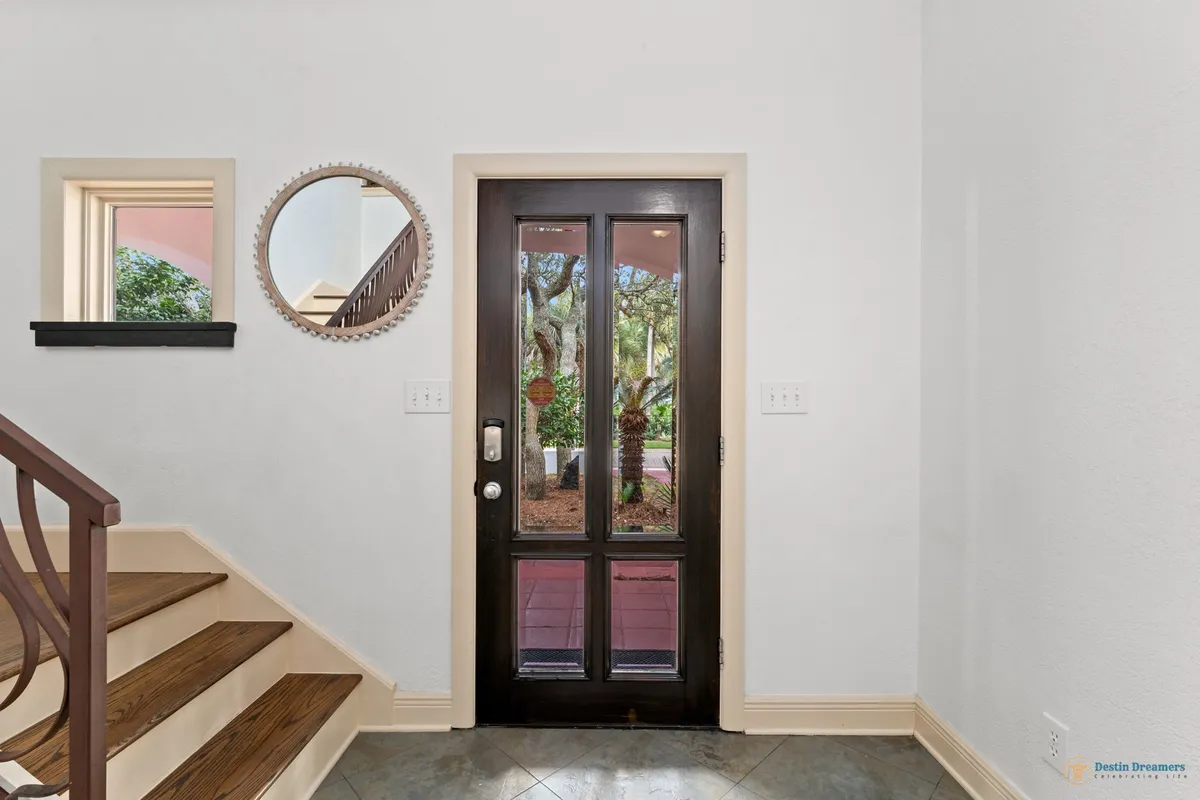A view from the interior showing a dark wooden door with glass panels that open to a lush garden, along with a wooden staircase and a decorative mirror reflecting light. The scene suggests easy access to outdoor areas and a fluid connection between inside and out.