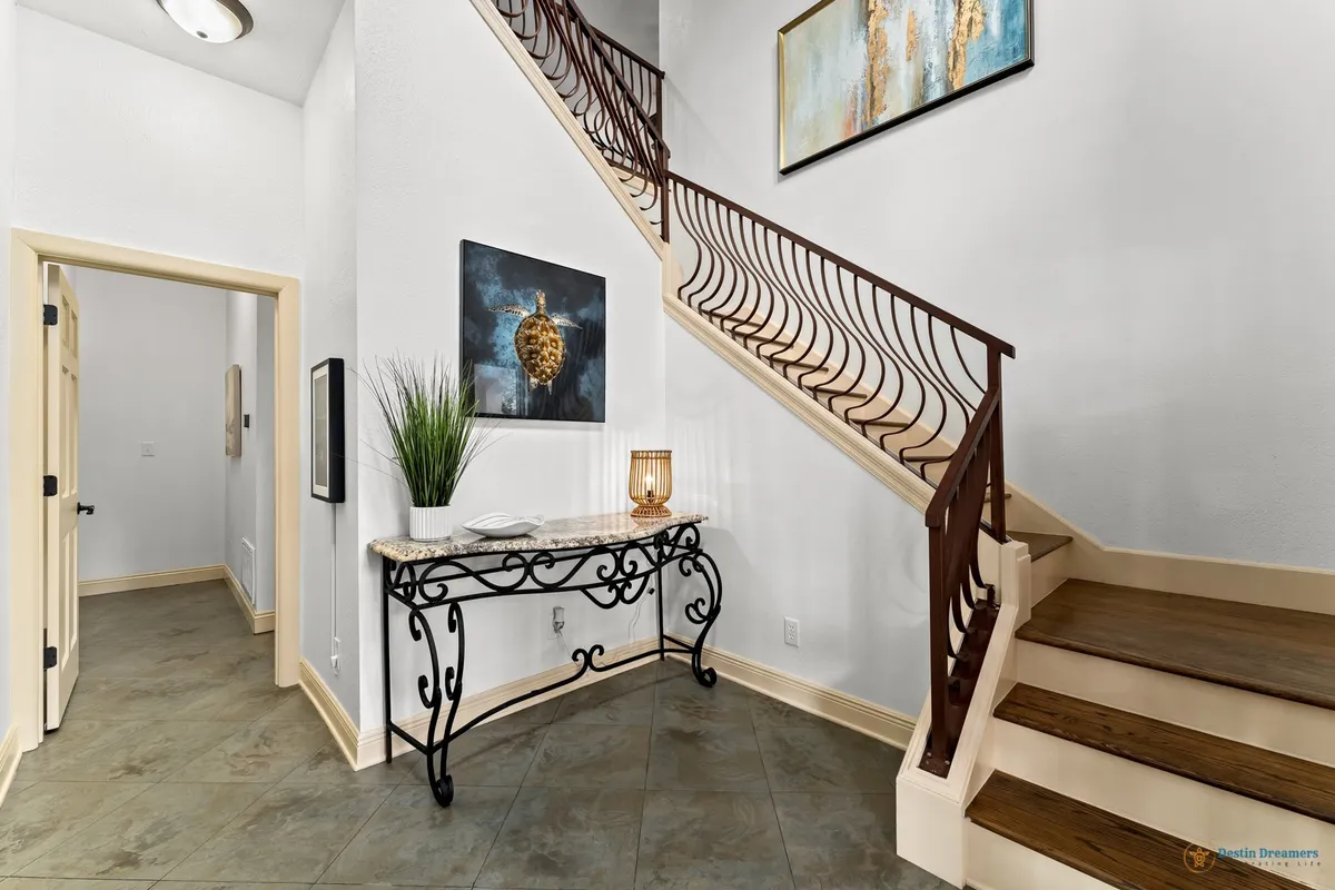 A welcoming entry hallway featuring an ornate wrought iron console table with a marble top, decorative wall art with a sea turtle motif, a wooden staircase with a decorative iron railing, and a potted plant. The mood is calm, elegant, and inviting—setting the tone for the rest of the home.