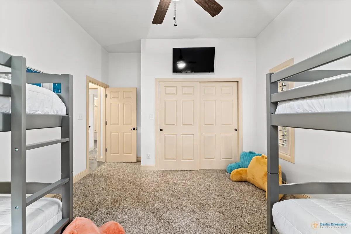 A cozy bunk room with two gray twin-over-twin bunk beds, a wall-mounted television above closet doors, plush toys in the corner, and carpeted flooring. The space is playful and inviting, designed for kids to relax, play games, and unwind.