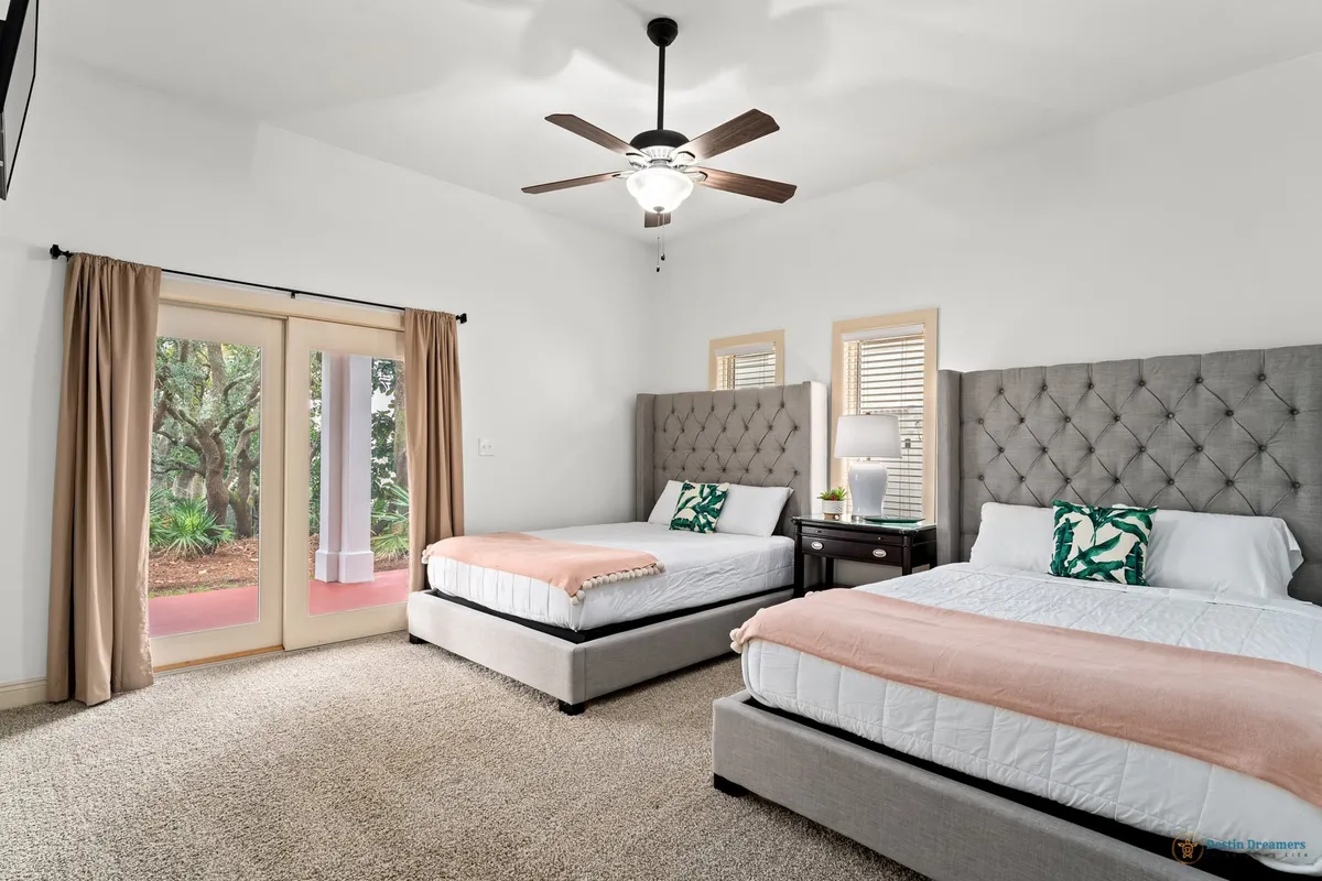 A spacious bedroom with two queen beds featuring tufted headboards, French doors opening to an outdoor area, a ceiling fan with a light fixture, and a nightstand with a lamp and decorative plants. The room is filled with natural light and evokes a sense of peaceful retreat.