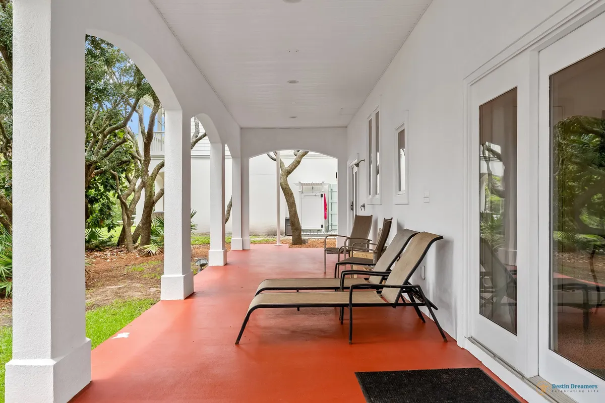 A peaceful exterior veranda lined with arched white columns, furnished with beige lounge chairs and a small table. The red painted concrete floor adds warmth, and the scene is surrounded by tranquil natural greenery, suggesting a restful outdoor spot for families.