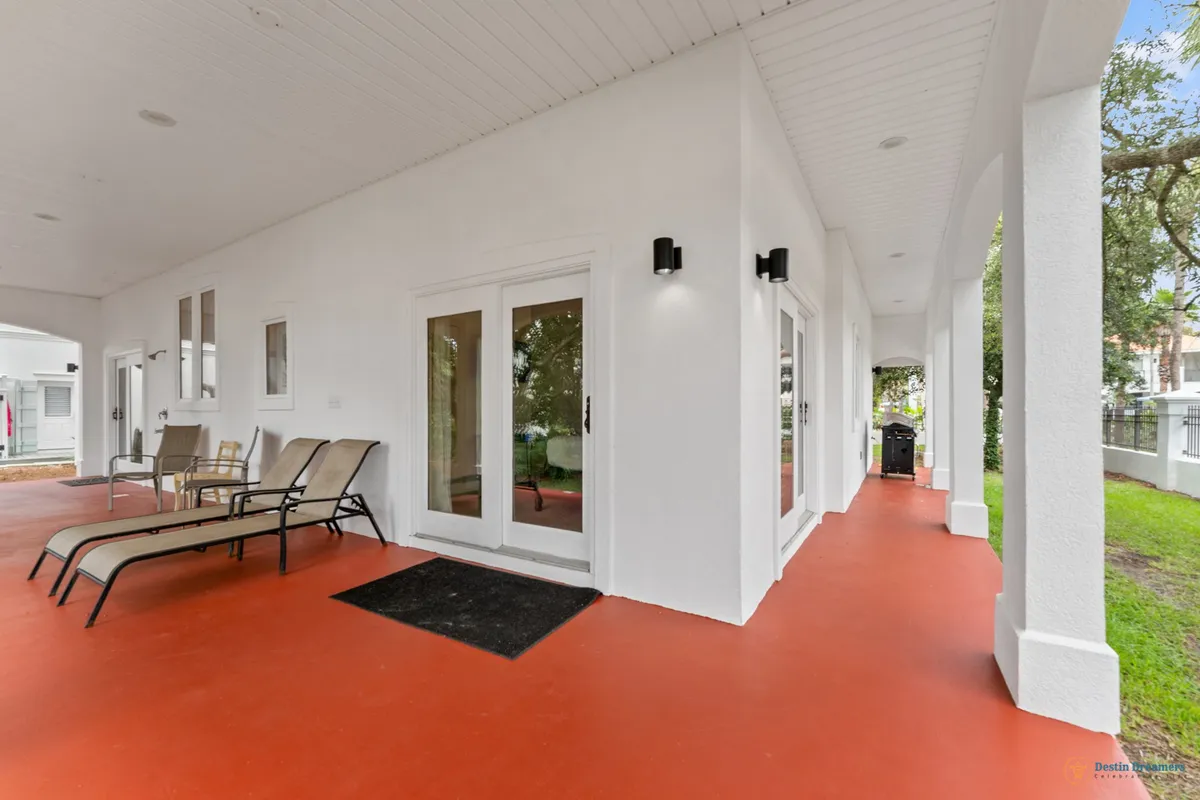A serene exterior veranda with white pillars and arches frames an inviting space. The terracotta-colored floor contrasts with the white walls, while lounge chairs offer a relaxing spot with easy access to indoor areas through double glass doors. The scene exudes privacy and calm, ideal for unwinding or socializing outdoors.