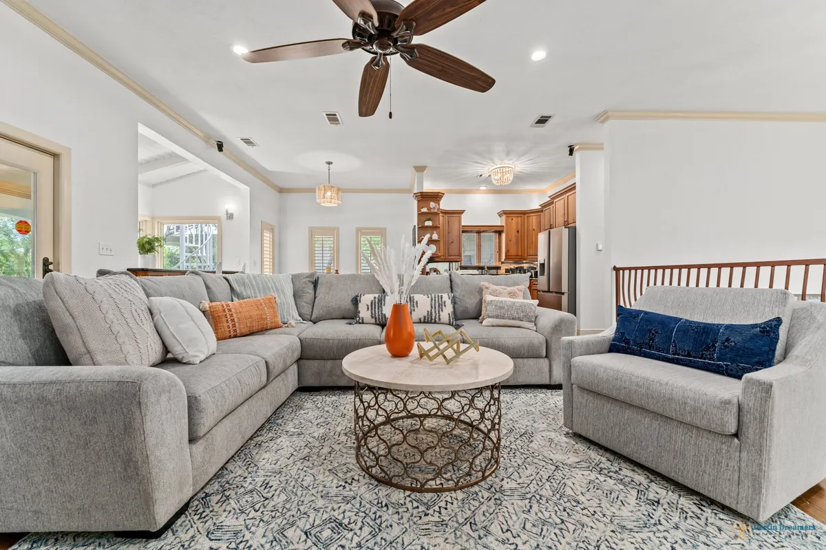 A spacious open-concept living area with a large grey sectional sofa accented by decorative pillows. A round coffee table with an intricate metal base sits in the center, while a ceiling fan with wooden blades hangs overhead. In the background, an open kitchen with wooden cabinetry is visible, reinforcing the sense of connectivity between living, dining, and kitchen spaces.