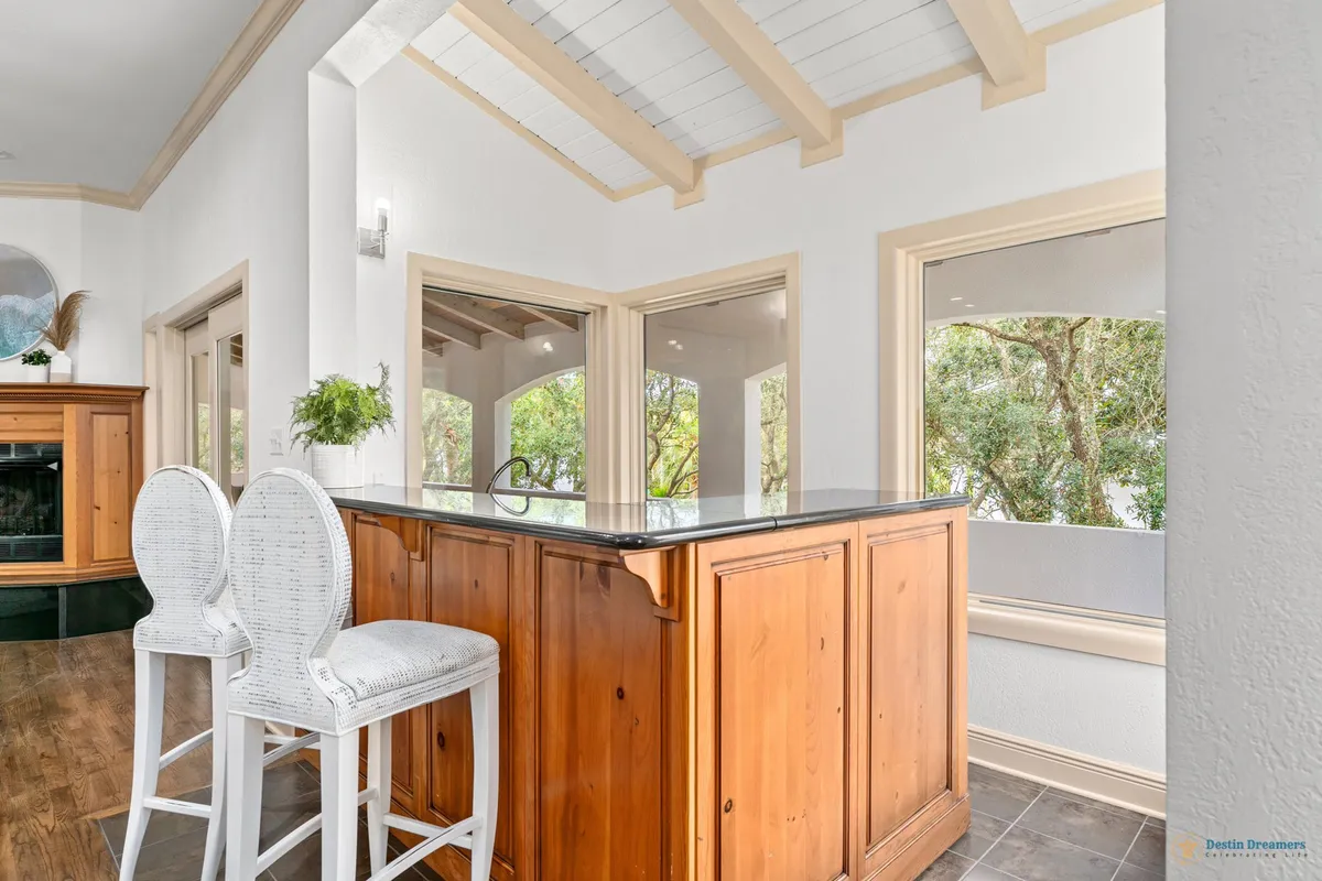 A bright, welcoming kitchen with a wooden island topped by a black countertop, surrounded by white high-back bar stools. Large windows fill the room with natural light and offer views of greenery outside. The vaulted ceiling with exposed beams adds architectural interest and a sense of spaciousness.