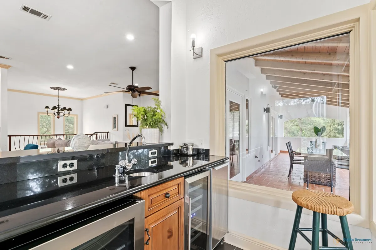 An open-concept living and dining area connects to a large window overlooking a covered balcony, with a black granite countertop and wine cooler in the foreground. The view toward the outdoor balcony area hints at the seamless flow between indoor comfort and outdoor al fresco dining, suggesting the convenience of preparing and enjoying meals while taking in Gulf breezes.