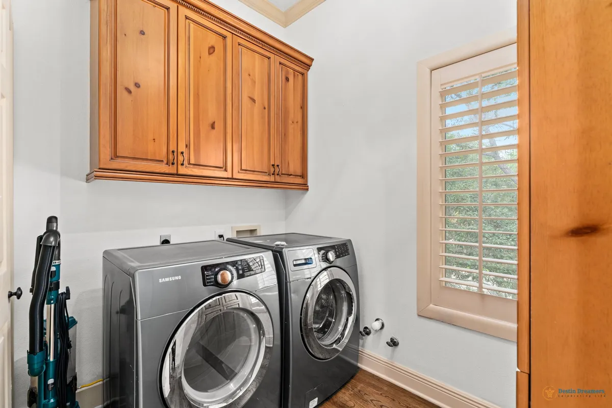 A practical laundry room featuring a front-loading washing machine and dryer, wooden upper cabinets for storage, and a window with plantation shutters that lets in natural light. The clean, organized layout supports easy vacation routines.