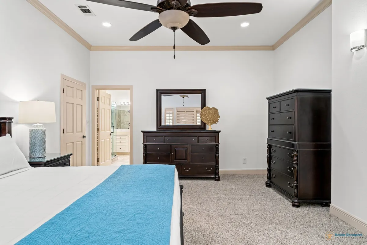 A spacious, tranquil master bedroom featuring a king-sized bed with a blue throw, complemented by dark wood furniture including a dresser with mirror and a tall chest of drawers. The room is filled with natural morning sunlight, enhancing the restful, serene atmosphere ideal for relaxation and privacy.