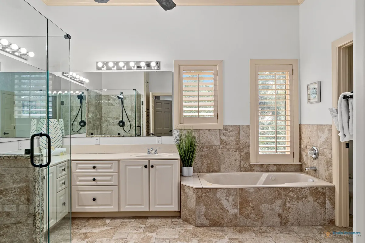 A spa-inspired bathroom with a built-in jetted bathtub surrounded by tile, a large glass-enclosed shower with black hardware, and a wide vanity with dual light fixtures. Plantation shutters allow gentle natural light to fill the space, creating a calm, rejuvenating ambiance.