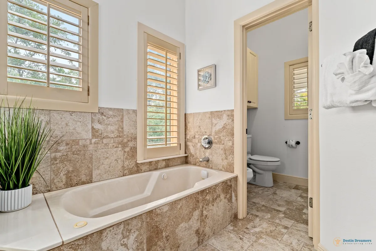 A soothing bathroom scene with a jetted bathtub, separate toilet area, decorative plant on the tub ledge, and windows with plantation shutters. The space is bright, clean, and invites quiet moments of relaxation and self-care.