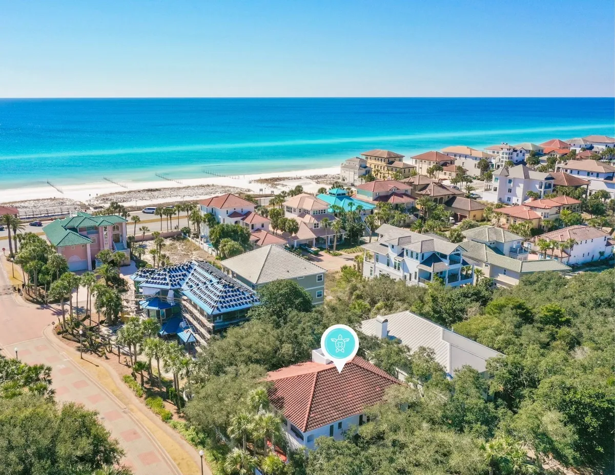 A breathtaking aerial view of Destin's turquoise Gulf waters and sugar-white sandy beach, bordered by a cluster of colorful upscale beach homes surrounded by lush green trees. The shoreline is inviting, with plenty of space for sunbathing, swimming, and family fun. The overall scene radiates a tranquil, upscale beachside retreat atmosphere.