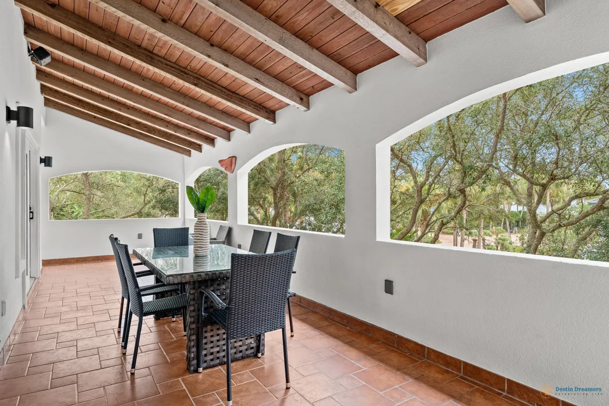 This image showcases a serene indoor-outdoor dining space, with a glass-top dining table and black wicker chairs set under a wooden beamed ceiling. Arched openings reveal lush green trees outside, and the terra-cotta tiled floor adds warmth. The view through the arches evokes a connection to the outdoors while being sheltered and comfortable.