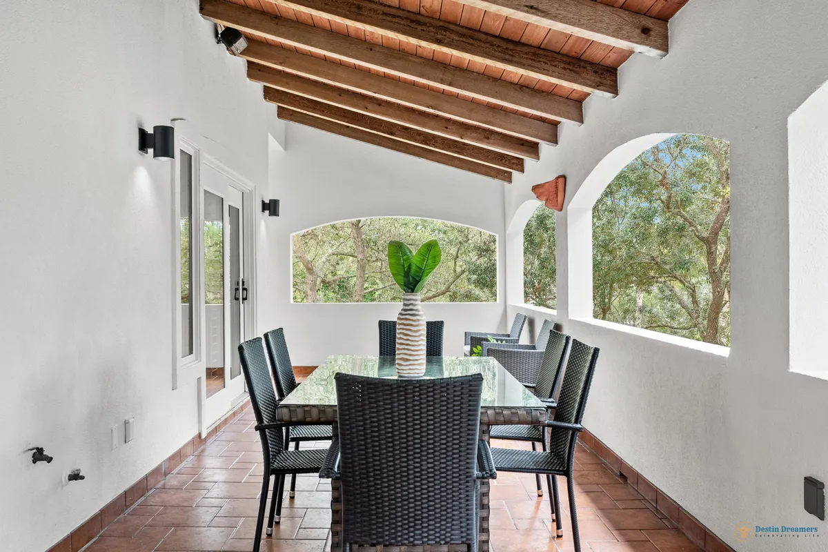 A light-filled dining area with arched open windows framing lush greenery outside, a glass dining table with wicker chairs, and a wooden beam ceiling. The decorative vase with green leaves as a centerpiece hints at a seamless connection to the natural world beyond, inviting guests to enjoy both indoor and outdoor experiences.