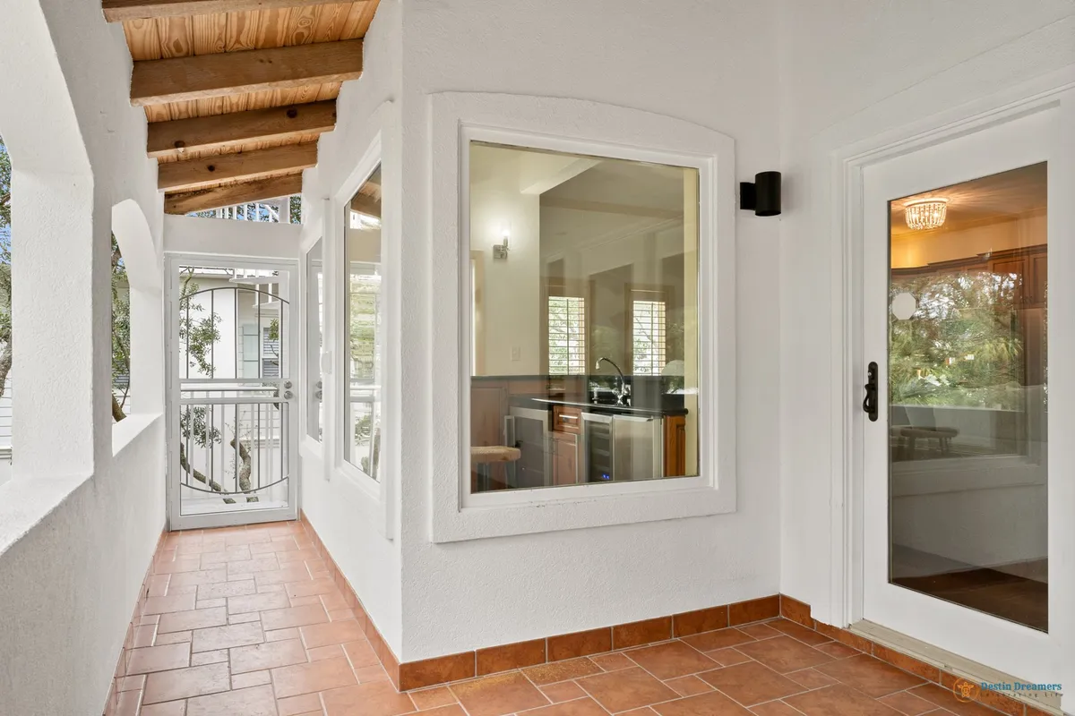 This image features arched openings along a spacious veranda with a large window providing a view into the kitchen, glass door with a modern handle, and exposed wooden ceiling beams. The vista through the arches hints at the greenery and peacefulness outside, while the veranda itself is shaded and inviting—perfect for relaxing with a pet.