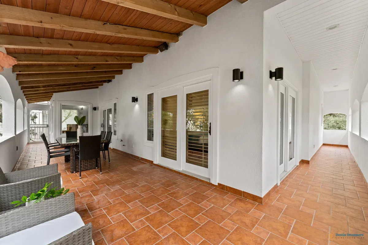 A long wrap-around veranda with terracotta tile flooring, a wooden beamed ceiling, an outdoor dining table with chairs, and French doors with white shutters opening to the interior. The space is bathed in warm, natural light and offers a tranquil view, inviting relaxation and social gatherings.