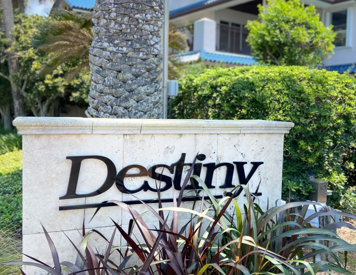 The welcoming entrance to the Destiny community, featuring a prominent 'Destiny' entrance sign set in a stone wall, landscaped with palms, and backed by residential buildings with blue roofs. The scene exudes a sense of exclusivity and tranquility, hinting at the upscale, gated environment inside.