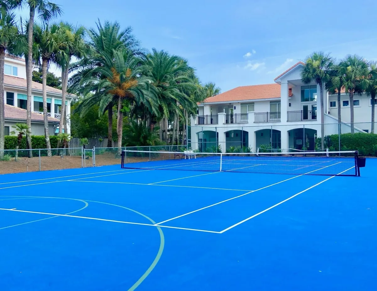 A bright blue multi-sport court, outfitted for tennis and basketball, sits surrounded by tall palm trees and a luxurious white building with terracotta roofing in the background. The vibrant court is designed for both active play and social gatherings, with a tropical, resort-like ambiance that signals access to premium community amenities.