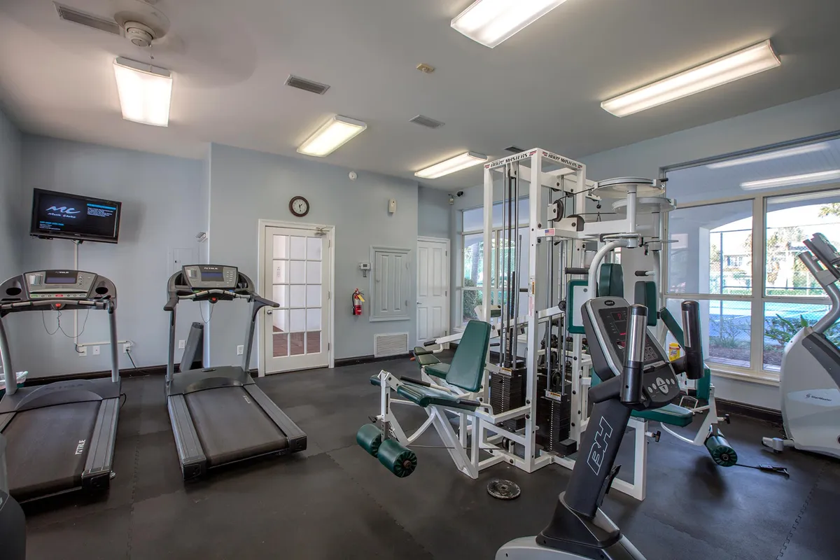 A well-equipped fitness center features treadmills, a multi-station gym machine, and a stationary bike, all bathed in natural light from large windows with views outside. The modern, airy space encourages health and wellness in a resort-like environment.